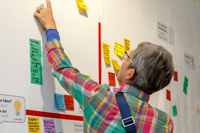 Fotografie - Person steht vor einem Whiteboard, auf dem Zettel mit Ideen zu sehen sind und klebt selbst einen Zettel auf